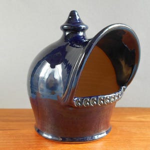 May include: A dark blue ceramic salt pig with a glossy finish. The salt pig has a rounded body, an open front, and a decorative rim. It features a small, pointed top and sits on a wooden surface.