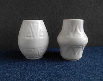 May include: Two white ceramic vases with geometric designs. The vase on the left has a more intricate design with triangles and squares. The vase on the right has a simpler design with curved lines.