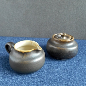 May include: A brown ceramic creamer and sugar bowl set. The creamer has a light brown rim and the sugar bowl has a lid.