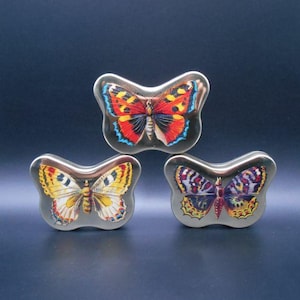 Pode incluir: Três latas decorativas em forma de borboleta. A lata superior apresenta uma borboleta com asas vermelhas, laranja e azuis. A lata inferior esquerda tem asas amarelas e brancas, e a lata inferior direita tem asas roxas e amarelas.