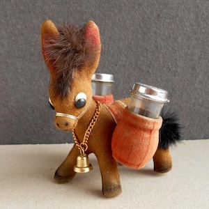 Könnte beinhalten: Ein braunes Plüsch-Esel-Salz- und Pfefferstreuer-Set. Der Esel trägt eine goldene Kette und eine Glocke. Die Salz- und Pfefferstreuer sind am Rücken des Esels befestigt.