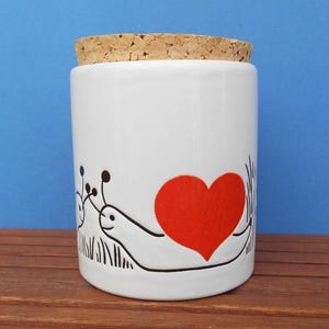 May include: White ceramic jar with a cork lid. The jar has a whimsical design of a snail with a red heart and black line art. The cylindrical jar sits on a wooden surface against a blue background.