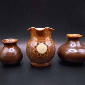 Può includere: Tre vasi in ceramica marrone, uno con un motivo floreale bianco e marrone. I vasi sono di diverse dimensioni e forme.
