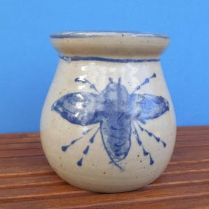 May include: A ceramic vase with a beige base and a blue bee design. The vase has a rounded body and a slightly flared rim. The bee is painted in a stylized manner, with detailed wings and legs. The background is a solid blue.
