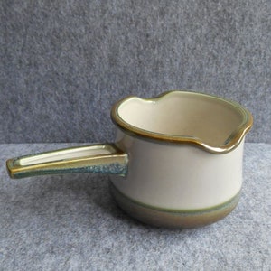 May include: A small, off-white ceramic saucepan with a handle. The rim and base have a brown and green trim. The handle is rectangular with a green and gold color. The interior is white.