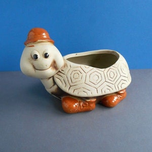 Może przedstawiać: Ceramiczna doniczka w kształcie uśmiechniętego żółwia z małym pomarańczowym kapeluszem. Skorupa żółwia ma geometryczny wzór, a jego stopy są również pomarańczowe. Doniczka jest przeznaczona do małych roślin lub kwiatów.