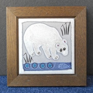 May include: A framed ceramic tile with a white polar bear illustration. The bear is walking on a blue and white patterned surface with a fish design. The tile is framed in a dark brown wood frame.