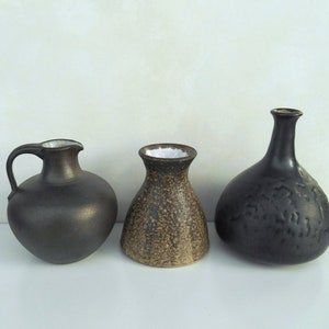 Puede incluir: Tres jarrones de cerámica de diferentes formas y tamaños. El jarrón de la izquierda es una jarra marrón oscura con asa. El jarrón del centro es un jarrón redondo más pequeño con un esmalte marrón moteado. El jarrón de la derecha es un jarrón redondo y alto con un esmalte marrón oscuro y una superficie texturizada.