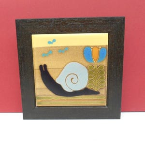 May include: Framed ceramic tile featuring a stylized snail with a light blue shell and black body. The artwork includes a blue and orange flower, green foliage, and small blue birds against a tan background. The frame is dark brown.