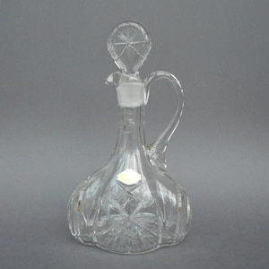 May include: Clear glass decanter with a starburst design on the base and stopper. The decanter has a handle and a spout for pouring. The stopper is a star shape. A small white label is on the front.