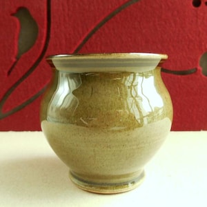 Peut inclure: Un pot en céramique vert olive fait à la main, avec un corps arrondi et un bord légèrement évasé. Le pot a une finition brillante et repose sur une petite base circulaire. Le fond présente une surface rouge avec un motif décoratif d'oiseau et floral.