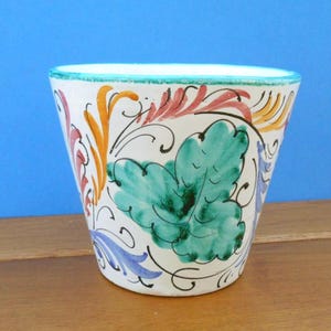 Puede incluir: Una maceta de cerámica blanca con un diseño pintado a mano. La maceta presenta un borde turquesa y está decorada con motivos florales y de hojas coloridos en tonos verdes, naranjas, rojos y azules. La maceta está sobre un fondo azul.