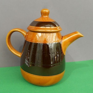 May include: A ceramic teapot with a lid, handle, and spout. The teapot is primarily brown and tan, with a dark brown band around the middle. The handle and spout are tan, and the lid has a small knob. The teapot has a classic, rounded shape.