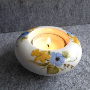 May include: A white ceramic candle holder with a lit tea light inside. The holder is decorated with hand-painted yellow and blue floral designs. The candle holder is round and has a diameter of approximately 10 cm.