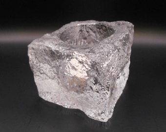 May include: A clear, textured glass candle holder with a square shape and a circular indentation for a candle. The glass has a rough, icy appearance, reflecting light and creating a shimmering effect. The candle holder is a decorative item.