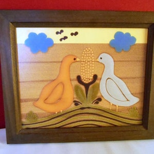 May include: A framed artwork featuring two birds, one orange and one white, perched on a brown and green landscape. The birds are facing each other and appear to be kissing. The background includes two blue clouds and a yellow sky.