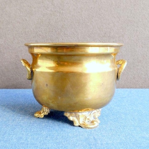 Könnte beinhalten: Ein kleiner, antiker Messingtopf mit zwei Griffen und drei verzierten Füßen. Der Topf hat einen abgerundeten Körper und einen breiten Rand. Das Messing hat eine warme, goldene Farbe. Der Topf wird wahrscheinlich zu dekorativen Zwecken verwendet.