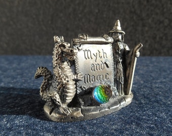 Puede incluir: Figurilla plateada con un dragón, un mago y un pergamino con las palabras "Myth and Magic". El dragón está a la izquierda, mientras que el mago sostiene un bastón. Una esfera iridiscente colorida está en la base.