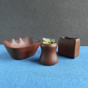 Pode incluir: Um conjunto de três itens de madeira: um cinzeiro pequeno, um isqueiro com a parte superior dourada e um pequeno recipiente quadrado. Os itens são feitos de madeira castanha escura e estão sobre uma superfície azul.