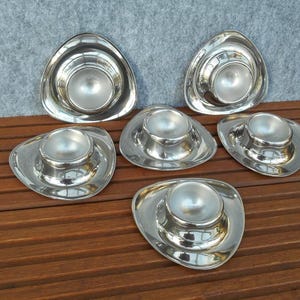 May include: A set of six silver-colored egg cups. Each cup has a rounded base and a central, egg-shaped holder. The cups are arranged on a wooden surface, reflecting light and creating a shiny appearance. The design is modern and minimalist.