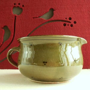 Pode incluir: Uma tigela de cerâmica artesanal com alça e bico, esmaltada em tons de verde oliva e castanho. A tigela tem formato arredondado e é decorada com pequenas manchas escuras. O fundo é um feltro vermelho com recortes de pássaros e flores.