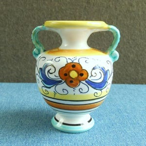 Op de afbeelding: Een kleine, decoratieve keramische vaas met een witte basis en een afgeronde body. De vaas heeft een gele rand en handvatten, met oranje, blauwe en gele bloemmotieven. De vaas is ongeveer 13 cm hoog.
