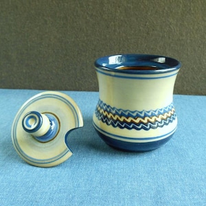 May include: A ceramic jar with a lid, featuring a cream-colored body with blue and brown zigzag and stripe designs. The rim and base are dark blue. The lid is off to the side, showing a matching pattern.