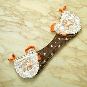 Puede incluir: Un guante de cocina acolchado con forma de pollo con un mango marrón con estampado floral. Las cabezas de los pollos son blancas con un estampado floral dorado y un corazón beige. Los detalles en naranja están en la cresta, el pico y las patas.