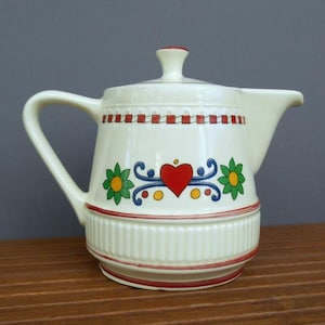 May include: A white ceramic teapot with a red, white, and blue floral design. The teapot has a heart-shaped design in the center of the floral pattern.