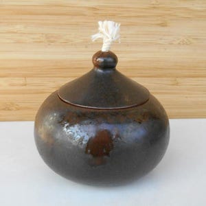 Può includere: Una lampada a olio in ceramica marrone scuro con uno stoppino bianco. La lampada ha un corpo arrotondato e un coperchio con un piccolo pomo in cima.