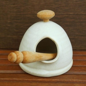 May include: A white ceramic birdhouse with a wooden perch and a wooden knob on top. The birdhouse has a rounded shape with a large opening for the birds to enter. The wooden perch extends from the opening.
