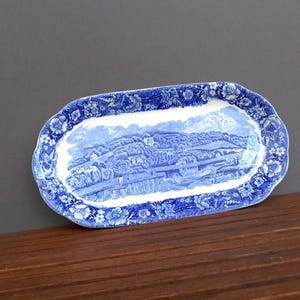 May include: Blue and white ceramic serving platter with a landscape scene. The rectangular platter has a scalloped edge with floral detailing. The center features a detailed depiction of a rural landscape.