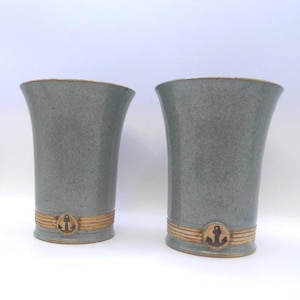 May include: Two grey ceramic vases with a nautical theme. Each vase has a flared top, a band of tan detail, and a brown anchor emblem. The vases are approximately 20 cm tall.