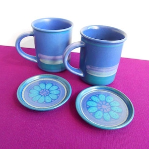 Könnte beinhalten: Zwei blaue Keramikbecher mit Henkeln und passenden Untersetzern. Die Becher haben einen blauen Sockel mit einem türkis-violetten Streifendesign. Die Untersetzer haben ein Blumenmuster in Blautönen.