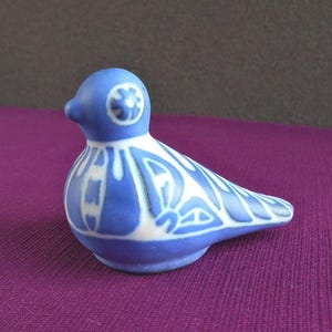 Może przedstawiać: Ceramiczna figurka ptaka w odcieniach niebieskiego i białego. Ptak ma zaokrąglony korpus i długi, spiczasty ogon. Ciało zdobi geometryczny wzór. Ptak siedzi na fioletowej powierzchni.