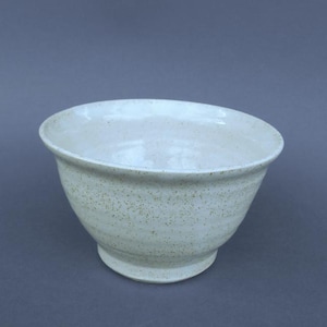 May include: A ceramic bowl with a wide, flared rim and a tapered base. The exterior and interior feature a light off-white glaze with a speckled texture throughout, showing small brown flecks embedded within the smooth, glossy finish of the vessel.