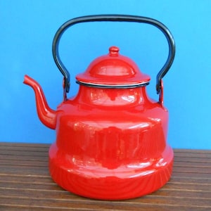 May include: A vibrant red enamel kettle with a black handle and spout. The kettle has a rounded body, a lid, and a black trim. The background is a solid blue color. The kettle is sitting on a wooden surface.