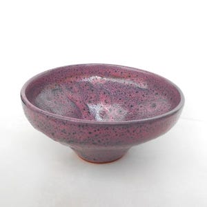 May include: A handmade ceramic bowl with a deep, rounded shape. The bowl features a speckled, glossy finish in shades of purple and black. The bowl has a footed base and is suitable for serving or display.