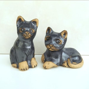 May include: Two ceramic cat figurines. One is sitting upright, the other is lying down. Both have black bodies with tan ears, paws, and facial features. The cats have detailed eyes and noses, and are on a white surface.