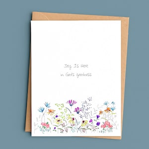 May include: A white greeting card with a watercolor floral design at the bottom and the text "Joy Is Here in God's goodness." The card is inside a brown envelope, set against a blue background.