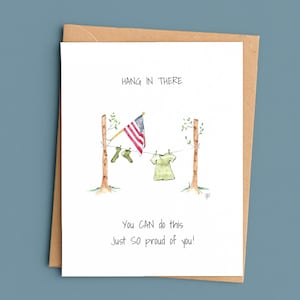 May include: A white greeting card with a watercolor illustration of a clothesline strung between two trees. The line holds two green socks and a green shirt, with an American flag hanging nearby. Text reads: HANG IN THERE, YOU CAN do this, Just SO proud of you!