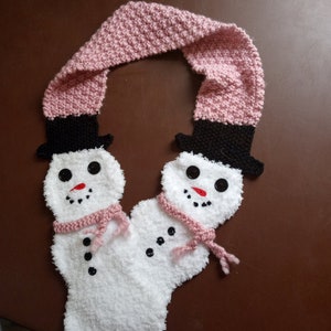 May include: A pink knitted scarf with two white snowman faces attached. The snowmen have black hats, black eyes, red noses, and black buttons.