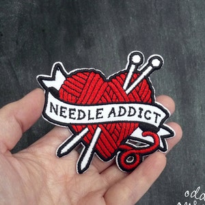 May include: Red heart-shaped embroidered patch with a ball of yarn and two knitting needles crossed inside. The banner below reads "NEEDLE ADDICT".