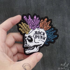 May include: An embroidered iron-on patch featuring a white skull with a mohawk of colourful crystals. The skull has the words "Rock Punk" written on its forehead.