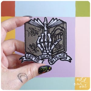 May include: Embroidered patch featuring a skeleton hand giving the middle finger, with an open book design. The patch includes the text "LET ME READ IN PEACE" on a banner. The patch is gray, black, and white.