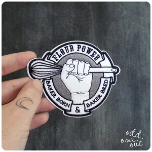 May include: A gray and white embroidered patch with a fist holding a whisk and the text "Flour Power Baker Born & Baker Bred".