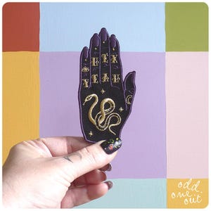 Puede incluir: Un parche bordado negro con forma de mano, con texto dorado y un diseño de serpiente dorada. El parche tiene uñas moradas y el texto "FIX YEAH". El fondo es un mosaico de cuadrados coloridos.
