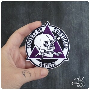 Può includere: Toppa ricamata bianca con un triangolo viola, uno scheletro con pennelli incrociati e il testo "Society of Tortured Artists" attorno al bordo.