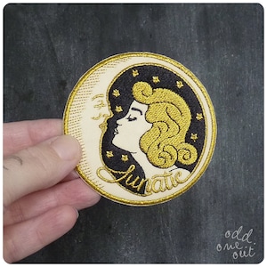May include: A gold and black embroidered patch featuring a woman's face with a crescent moon behind her. The text "lunatic" is embroidered below the woman's face.