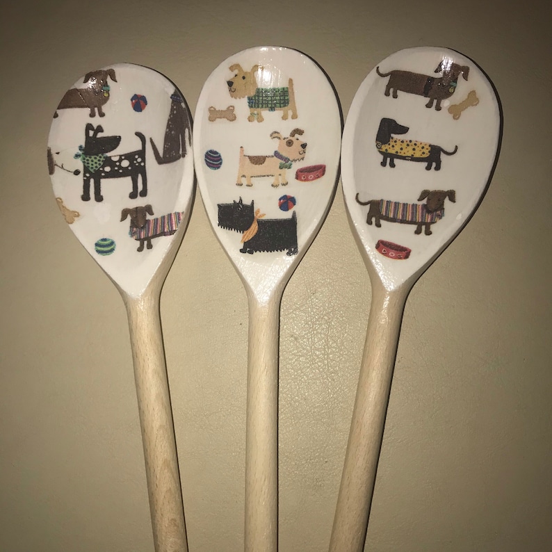 May include: Three wooden spoons with a white background and colorful dog illustrations. The dogs are wearing clothes and have different breeds. The spoons are a great gift for dog lovers.
